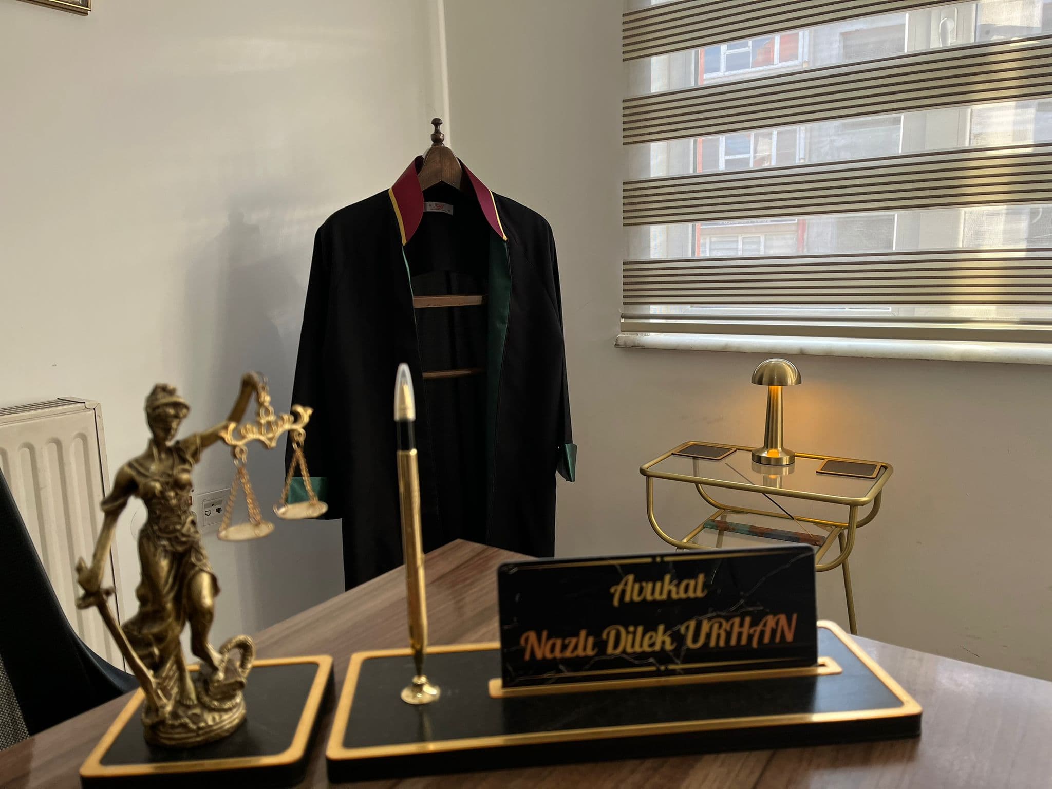 Burhaniye’deki Avukat Nazlı Dilek Urhan hukuk bürosundan çalışma masası ve iç mekan; avukatlık ruhsatı, cübbe ve adalet heykeli ile profesyonel danışmanlık alanı.